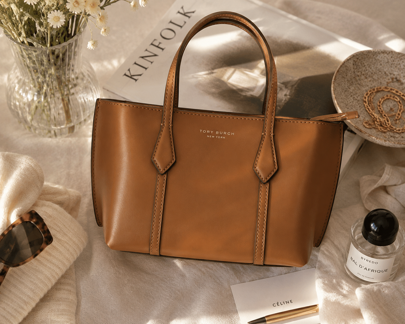 A tan Tory Burch leather tote styled with a Kinfolk magazine, sunglasses and perfume on cream linen, for SIGNVM